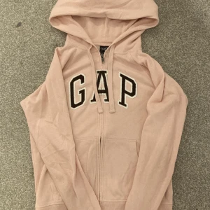 GAP zip upp hoodie - En ljusrosa GAP zip upp hoodie med bruna bokstäver, köpt secondhand men har själv aldrig använt den. Varan är i bra skick och ser knappt använd ut. Skriv gärna vid funderingar!!!