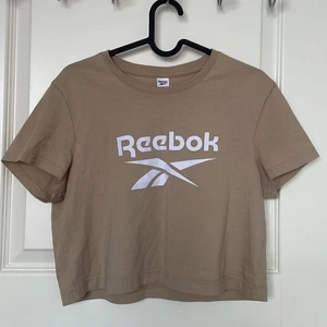 Reebok T-Shirt Storlek S/M - T-Shirt i jättebra skick, färgen är beige och texten vit, köpt från Stadium men märket är Reebok. Tröjan är i storlek M men den är ganska croppad så det passar personer med S också.