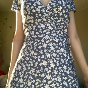 Blå omlottklänningen  - En blå omlottklänningen med vita blommor. Använd ca. 15 gånger men den ser helt ny ut. Den har tyvärr blivit lite för kort på mig.