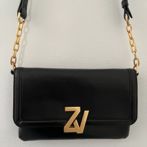 Zadig & Voltaire Clutch - Stilren Zadig & Voltaire Clutch i svart  läder med guld initialer. Dragkedjefack på insidan Justerbara remmar med gulddetaljer.    