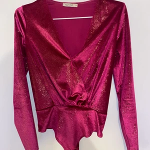 Glittrig lila/rosa body -  En lila/rosa, glittrig, omlott string body i storlek XS. Jätte skönt material men jag har inte haft användning av den mer än en gång. Passar till fest men även tex jul. Orginalpris 299. Kan fixa bilder hur den sitter på. Från NLYone