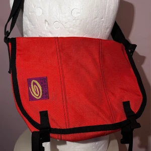 TIMBUK2 VÄSKA, SMALL. RÖD. FINT SKICK. RETRO, VINTAGE, 90-TAL. - Röd retro/vintage Timbuk2 väska.  Storlek: Small, ca 36 cm bred, 25 cm hög, 16 djup.  Fint skick, dock saknas reflexer och midjerem - se bilder för egen bedömning.   Har flera små fack.    Trendig 1990-tals budväska.