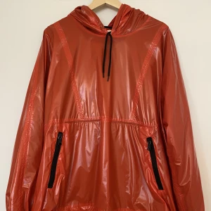 Our Legacy Festival Parka, garbage red - Röd parka från Our legacy i ett glansigt polyester-tyg. Svart mesh-foder. Storlek 48/Medium och passformen är boxig. Se mått nedan,  Längd: 71 cm Axelbredd: 61 cm Ärmlängd: 58 cm  Skick: 8/10 (nyskick med undantag för en lite svart fläck på luva, se bild)