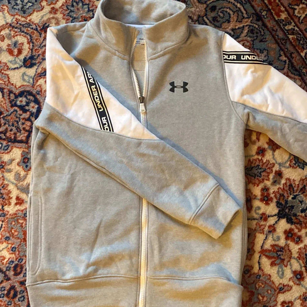 Långärmad Under Armour grå tröja med vita detaljer & dragkedja. Använd en gång. Skön insida i form av tunn fleece. Storlek anges ej, men    för barn ca 150 cm. Tröjor & Koftor.