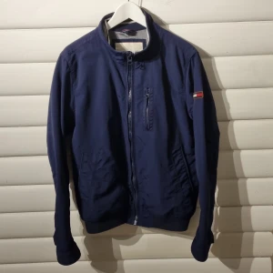 Tommy Hilfiger Jacka - Tunnare typ av jacka i bra skick för att vara från runt sent 00-tal, inga hål eller liknande men lite löstråd.  Är själv 180cm 70kg, jämför gärna med sista bilden.  Mått: Axelbredd 46cm. Total höjd 66cm. Armlängd 66cm.