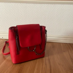 Röd ryggsäck - Liten, enkel och stilren väska med justerbara axelband och svarta kedje-detaljer. Även kulor på undersidan för att minimera att den ska bli sliten snabbare som även gör den väldigt stabil. Den är rymlig och lätt. Rätt så använd men det syns inte alls.