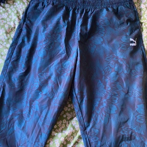 Puma regnbyxor - Ett par oanvända regnbyxor/vindbyxor från Puma. Storlek XXL, svarta med blommigt mönster i teal. 