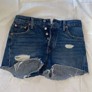 Ett par indigoblåa jeansshorts från Levis i modellen 501. Shortsen har lite fransar och hål samt knappar vid gylfen. Shortsen är i storlek W24. ✨🤍