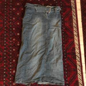 Maxikjol av jeanstyg. Med ett bälte och en slits där bak. 85 cm i midjan, 97 cm lång