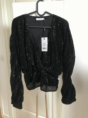 Svart långärmad blus från NAKD - Säljer en svart långärmad blus med paljetter och ballongärmar i storlek 36 🤩 Plagget är oanvänt med lappar på från NAKD 🏷