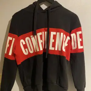 En svart röd hoodie med texten ”confidence” tvärs över. Använt men bra skick.🎀