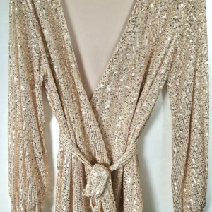 Paljettklänning Omlott i Guld - Oanvänd glamorös guldig paljettklänning i omlott med band som man knyter i midjan. Fin både på sommaren liksom till nyår. Helt ny!