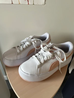 Vit/rosa Puma skor - Ett par vita Puma skor med rosa/aprikos färgade inslag använda endast 2 gånger. Skorna är i bra skick. Sulan är ganska hög och i storlek 39. Det finns ett litet märke längst fram på skon men annars i bra skick. 
