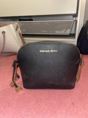 Svart micheal kors axelremsväska  - En svart axelremsväska från Micheal kors. Svart väska med guldkedja + läder band. Köpte för 1200kr. Säljer för 700kr. Först till kvarn. Möts i Stockholm eller fraktar