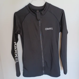 Barrel rashguard  - Barrel rashguard eller träningstopp med dragkedja. Svart med vit logga och text. Endast använt ett par gånger, perfekt skick. Kan användas som solskydd/cover up vid surfing etc eller som träningströja. Strl M men passar som S pga asiatisk sizing.