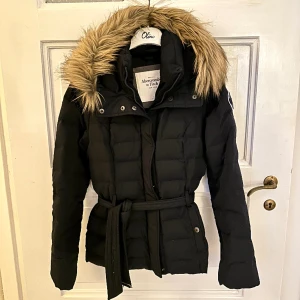 Dunjacka från Abercrombie & Fitch - Mörkblå dunjacka med markerad midja från Abercrombie & Fitch. Fejkpäls på luvan som är avtagbar. Jackan är i storlek M men passar främst S
