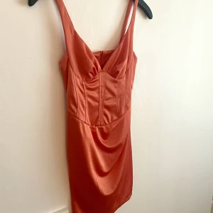 Orange/korall kort klänning - Orange miniklänning i satin med smala axelband och korsett med kupor från Asos. Klänningen drar in midjan och lyfter brösten. Aldrig använd med lapp kvar. Otroligt fin men säljer pga klänningen är för kort för mig (jag är 176). Nypris var 500 kr. 