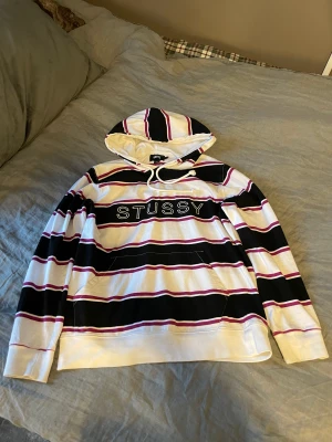 Stüssy hoodie  - Stüssy hoodie med ränder i vit, svart och lila/röd. Bra skick, köpt second hand därav en liten fläck där bak men inget som stör. Snabb leverans, fler bilder finns 