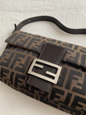 Fendi Baguette  - FENDI Zucca Baguette Bag  - Silverfärgade beslag - Enkel platt justerbar axelrem - Bruna läderdetaljer - Brunt foder och en ficka med dragkedja på innerväggen  Övergripande skick: Mycket bra. Utvändigt skick: Mycket bra. Lätt slitage och märken vid läderd