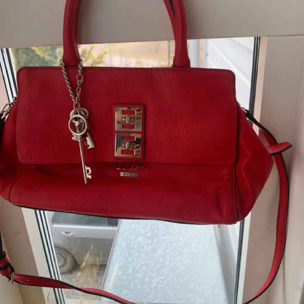 Röd guess handväska, långt band finns, ”keychain” hänger på ena handtaget 3 innefickor, 1 bak och 1 fram Silver detaljer Dust bag medföljer. Väskor.