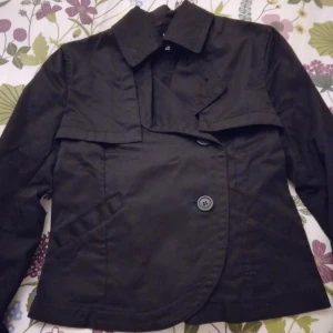 Kort svart vårjacka / sommarjacka. Strlk: XS/32. Fint skick. - Kort svart vårjacka / sommarjacka.   Strlk: XS/32. Material: Bomull. Ofodrad. Stängs med knappar. Har 2 fickor framtill. Inga defekter. FRAKT TILLKOMMER.