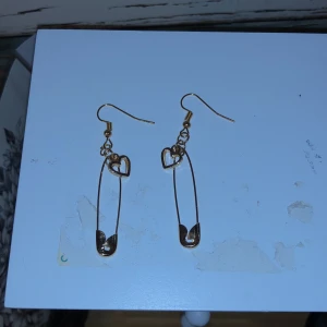 Örhängen Guldiga Säkerhetsnålar - Ett par fina örhängen med guldiga krokar och guldiga säkerhetsnålar med ett hjärta på. Dom är ej nickelfria, äkta guld eller rostfria