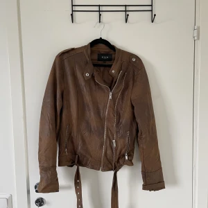 Läderimitationsjacka - Läderimitationsjacka brun, i gott skick. Skärp och fickor med dragkedjor!