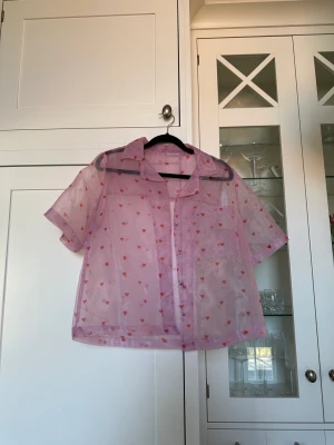 Organza Skjorta Hjärtmönster Skinnydip - Väldigt söt kortärmad skjorta i organzamaterial med hjärtmönster. Från märket Skinnydip, köpt på asos. Använd ett fåtal gånger. Lite boxig, oversize och croppad i modellen. Lappen i kragen är bortklippt då den syntes igenom tyget. Nypris 389 kr. 