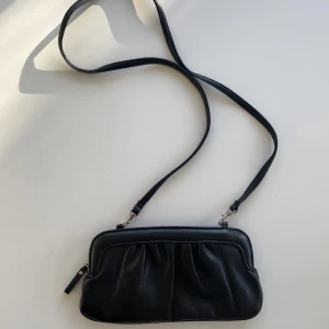 Söt svart väska - Vintageväska i svart i fint skick. Använd som clutch eller över axeln. Känns och ser ut som mjukt skinn men kan inte garantera. Längd ca 27 cm Höjd ca 14 cm.