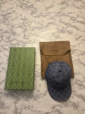 Blå denimkeps från Gucci - Säljer en snygg blå keps från Gucci i denim med det klassiska GG-mönstret över hela kepsen. Kepsen har böjd skärm och justerbar rem bak. Kommer med originalförpackning och dustbag.