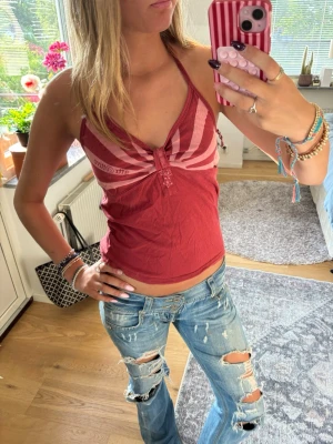 Röd halterneck linnetopp med mönster - Snygg röd linnetopp med halterneck och vita ränder upptill. Toppen har en knytning framtill och är ärmlös, perfekt för en trendig och avslappnad look. Materialet känns lätt och passar bra till jeans eller kjol.