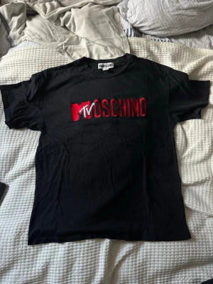Svart MTV x Moschino t-shirt - Snygg svart t-shirt från H&MOSCHINO med stor MTV-logga och MOSCHINO-text i rött och vitt på bröstet. Klassisk passform och rund hals. Perfekt statement-plagg för dig som gillar streetwear och coola samarbeten.