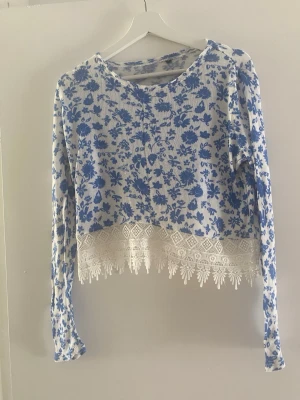 Blommig blå och vit croppad topp - Söt croppad topp med blått blommönster på vit botten och lång ärm. Nederkanten har en bred spetsdetalj. Perfekt för dig som gillar en trendig stil.