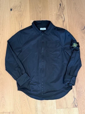 Svart overshirt från Stone Island - Snygg svart overshirt från Stone Island med dragkedja framtill, bröstficka och klassisk logotyp-patch på vänster ärm. Overshirten har en stilren design med krage och långa ärmar, tillverkad i ett lätt och slitstarkt material.