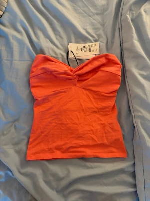Korallfärgad bandeau tubtopp - Säljer en snygg korallfärgad bandeau tubtopp med twistad detalj framtill. Toppen är axelbandslös och har en tight passform, perfekt för sommaren. 