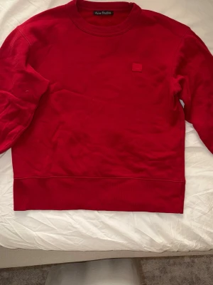 Röd sweatshirt från Acne Studios - Säljer en stilren röd sweatshirt från Acne Studios med klassisk rund hals och diskret Acne-logga på bröstet. Tröjan har ribbade muddar vid ärmslut och nederkant. Perfekt för dig som gillar minimalistisk design och vill ha något bekvämt och snyggt.
