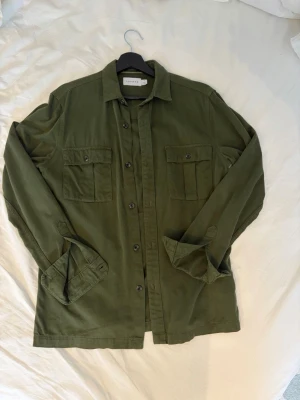 Olivgrön overshirt från Topman - Stilren olivgrön overshirt från Topman med klassisk krage, knäppning framtill och två bröstfickor med lock och knapp. Tillverkad i ett kraftigare bomullstyg som ger en avslappnad men snygg look. Perfekt att bära öppen över en t-shirt.