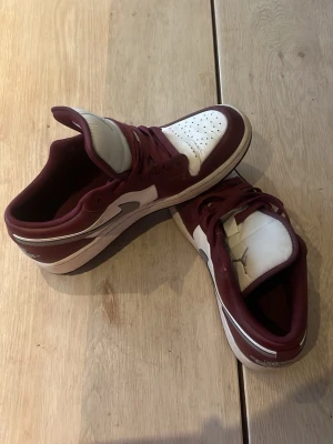 Vinröda och vita låga sneakers jordans  - Snygga sneakers i vinrött och vitt med klassisk låg siluett. Skorna har perforerad tåbox, matchande vinröda skosnören och kontrasterande detaljer. Ovandelen verkar vara i läder eller syntetmaterial och sulan är vit med vinröda inslag.