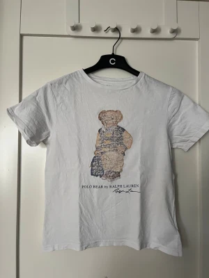 Vit Polo Bear t-shirt från Ralph Lauren - Vit t-shirt från Ralph Lauren med det ikoniska Polo Bear-motivet på bröstet. Klassisk rund hals och korta ärmar. Tillverkad i mjuk bomull för en skön känsla. Perfekt för en avslappnad och stilren look.