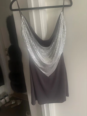 Kort svart klänning med silverdetaljer - Snygg kort svart klänning med tunna axelband och en draperad framsida täckt av glittrande silverpaljetter. Klänningen har en lös passform och ett elegant fall både fram och bak. Perfekt för dig som vill sticka ut med en trendig look.
