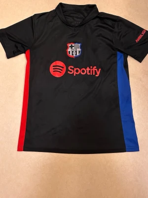 Svart FC Barcelona matchtröja - Säljer en svart FC Barcelona matchtröja med klubbmärke och Spotify-logga på bröstet. Tröjan har röda och blå sidopaneler samt korta ärmar. Perfekt för dig som älskar fotboll och vill stötta Barca på planen eller läktaren.