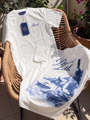 Vit t-shirt med blått tryck - Säljer en vit t-shirt med blått tryck på ryggen som föreställer vinflaska, glas och havsmotiv. Framtill står det 'Mariel' broderat i blått. T-shirten har rund hals och korta ärmar, perfekt för en avslappnad stil.