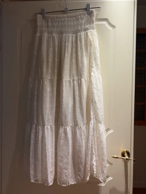 Vit långkjol  - Luftig vit långkjol från Gina Tricot med smock i midjan och volangdetaljer. Kjolen har ett diskret spetsmönster och är perfekt för varma dagar. Passar dig som gillar en romantisk och bohemisk stil.