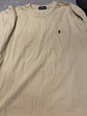 Beige t-shirt från Polo Ralph Lauren - Snygg beige t-shirt från Polo Ralph Lauren med klassisk rund hals och den ikoniska lilla loggan broderad på bröstet. Tillverkad i mjuk bomull som känns skön mot huden. Perfekt basplagg för en clean och stilren look.