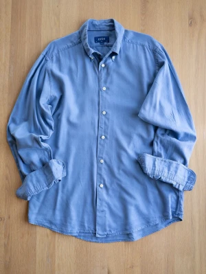 Blå skjorta från Eton i bomull, size L - Säljer en klassisk blå skjorta från Eton med knappar framtill och lång ärm. Skjortan har en stilren krage och är tillverkad i mjuk bomull, perfekt för en clean och avslappnad look.
