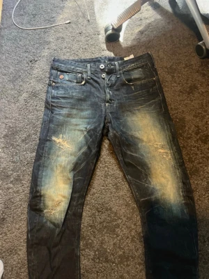 G-star jeans  - Säljer ett par mörkblå jeans med tydliga slitningar och ljusa partier på låren och knäna. Jeansen har fem fickor, knappgylf och en smal passform. Perfekta för en avslappnad och trendig look.