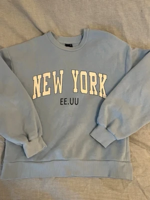 Ljusblå sweatshirt med New York-tryck - Ljusblå sweatshirt med stort vitt tryck där det står 'NEW YORK'. Tröjan har rund hals, ribbade muddar vid ärmslut och nederkant samt en avslappnad passform. 