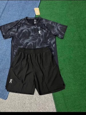 Svart träningsset från On - Snyggt träningsset från On med en svart t-shirt i abstrakt mönster och matchande svarta shorts. T-shirten har rund hals och korta ärmar, medan shortsen har elastisk midja och logga på benet. Perfekt för gymmet eller löpning.