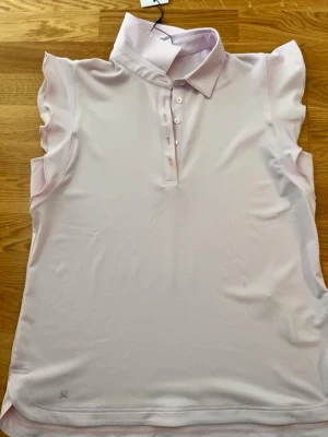 Ljusrosa ärmlös polotopp - Supersnygg ljusrosa ärmlös polotopp från Daily Sports med krage och knappar framtill. Toppen har volangdetaljer vid axlarna och är tillverkad i ett mjukt, stretchigt syntetmaterial som passar perfekt för sportiga aktiviteter.