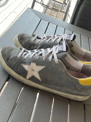 Grå GGDB sneakers med stjärna - Säljer ett par grå sneakers från GGDB med vit stjärna på sidan och gula detaljer vid hälen. Skorna har vita snören och en klassisk låg siluett. Ovandelen är i mocka och sulan är vit. Perfekta för dig som gillar streetwear och vill ha något unikt.